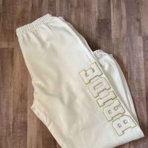 May include: White sweatpants with a gold embroidered "BIGGIE" logo on the leg.