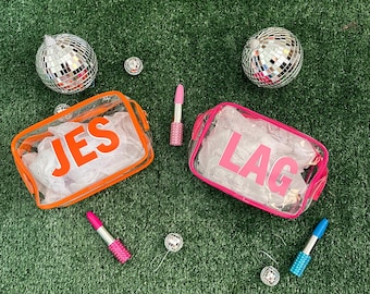 May include: Two clear plastic zippered pouches with orange and pink trim. The pouches have the names "JES" and "LAG" printed in white letters. The pouches are on a green artificial grass surface with silver disco balls and pink and blue lipstick tubes.
