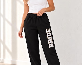 May include: Black sweatpants featuring the word BRIDE printed in white block letters with a gold outline on the left leg. The sweatpants have an elastic waistband and elastic cuffs at the ankles, paired with a simple white tank top and white sneakers.
