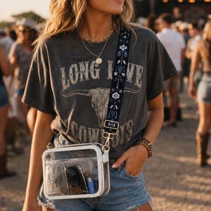 May include: A charcoal gray short-sleeve t-shirt featuring a graphic of a bull skull and the text LONG LIVE COWBOYS. The shirt is paired with a clear rectangular crossbody bag that has a dark blue patterned strap with white floral accents.