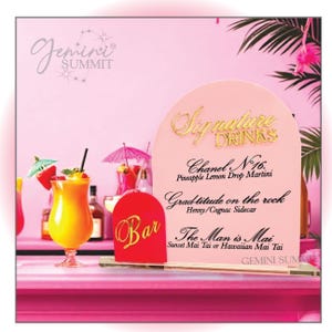 May include: A pink and white sign with gold lettering that reads "Signature Drinks". The sign lists three cocktails: "Chanel N°16. Pineapple Lemon Drop Martini", "Grad titude on the rock Henny/Cognac Sidecar", and "The Man is Mai Sunset Mai Tai or Hawaiian Mai Tai". The sign is on a bar with a red and gold "Bar" sign.