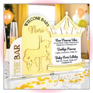 May include: A gold-colored acrylic bar sign with a carousel design, featuring the text "WELCOME BABY Nori" and drink names. The sign includes a carousel horse silhouette and the words "THE BAR" on the side. The sign is decorated with gold confetti.
