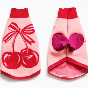 May include: Pink dog sweater with a red cherry design and bow. The sweater has a red collar and trim. The back of the sweater has two openings for the dog's legs, with pink and red accents.