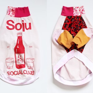 Puede incluir: Camiseta para perro con cuerpo blanco y gráfico rojo de "Soju". El diseño incluye una botella de soju roja, vasos y la frase "Social Club". El cuello es rosa con estampado floral.