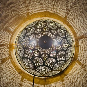 May include: A round, dark metal wall lamp with a detailed, perforated design. The lamp casts intricate patterns of light and shadow on the surrounding wall. The lamp is mounted on a gold-colored frame and has a black power cord.