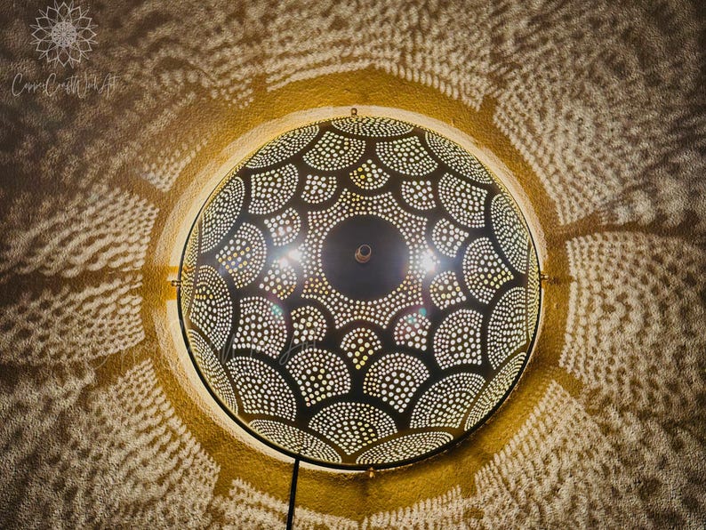 May include: A round, dark metal wall lamp with a detailed, perforated design. The lamp casts intricate patterns of light and shadow on the surrounding wall. The lamp is mounted on a gold-colored frame and has a black power cord.