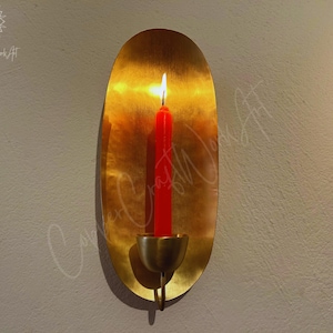 May include: A gold oval wall sconce with a candle holder. A red candle is lit inside the holder.