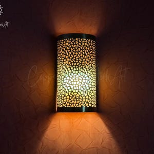 Peut inclure: Applique murale cylindrique avec un abat-jour en métal perforé, projetant une lumière chaude et modelée. L'applique est fixée sur un mur texturé et sombre. Le design présente un motif en mosaïque, créant un effet d'éclairage décoratif.