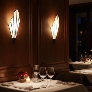 May include: Two wall-mounted sconces with a fan-shaped, geometric design and white frosted glass panels. The fixtures emit a warm, soft glow against dark wood paneled walls, illuminating a dining table set with white linens and glassware.