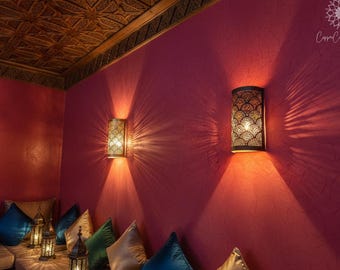 May include: Two cylindrical wall sconces with intricate cut-out patterns mounted on a textured magenta wall. The lights cast bright, starburst-shaped shadows against the wall. Below, several decorative pillows in blue, green, and gold are visible.