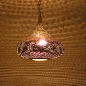 Peut inclure: Un luminaire suspendu décoratif avec un abat-jour en métal perforé. Le luminaire est de couleur or chaud et est suspendu à une chaîne. L'abat-jour est parsemé de petits trous, permettant à la lumière de filtrer. La lumière est allumée.