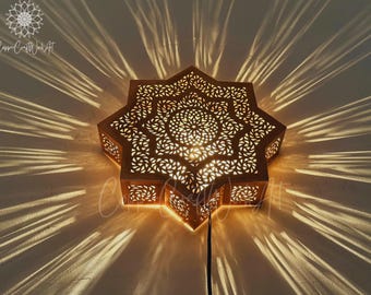 Può includere: Applique da parete decorativo a forma di stella con intricati motivi traforati. L'apparecchio di illuminazione dai toni caldi proietta un motivo di luce e ombra sulla parete. L'apparecchio è in metallo dai toni caldi e ha un design floreale centrale.