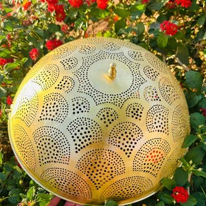 May include: A golden, dome-shaped metal lamp shade with intricate cut-out patterns. The shade has a small knob on top and is surrounded by red flowers and green foliage. The design features a series of semi-circles and circular patterns.