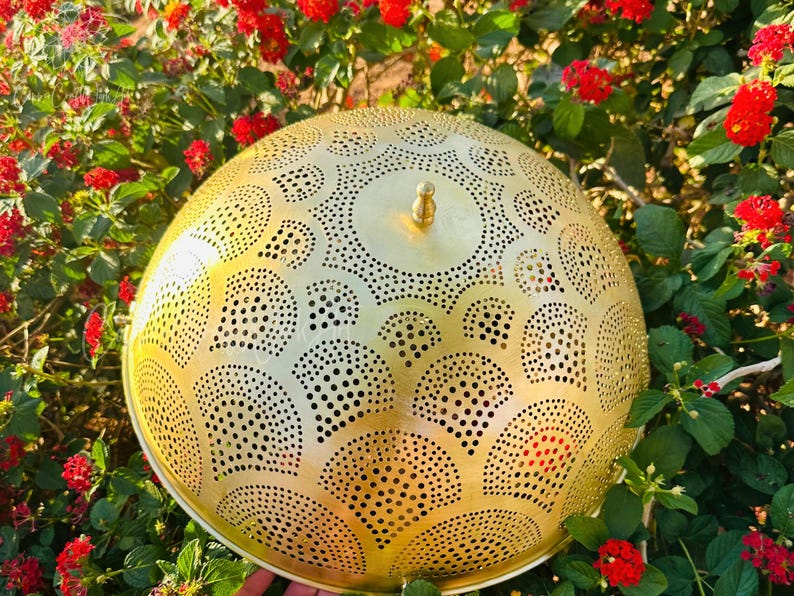 May include: A golden, dome-shaped metal lamp shade with intricate cut-out patterns. The shade has a small knob on top and is surrounded by red flowers and green foliage. The design features a series of semi-circles and circular patterns.