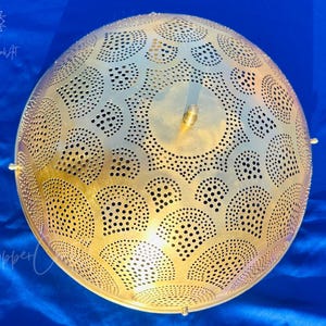 May include: A golden, spherical pendant light with intricate cut-out patterns. The design features a series of semi-circles and circular perforations. The light fixture is suspended and has a brass finish. The background is a deep blue fabric.