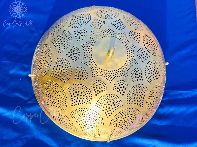 May include: A golden, spherical pendant light with intricate cut-out patterns. The design features a series of semi-circles and circular perforations. The light fixture is suspended and has a brass finish. The background is a deep blue fabric.