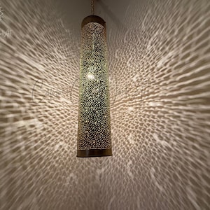 May include: A gold metal hanging lamp with a cylindrical shade featuring a decorative pattern of small holes. The lamp casts a patterned shadow on the wall.