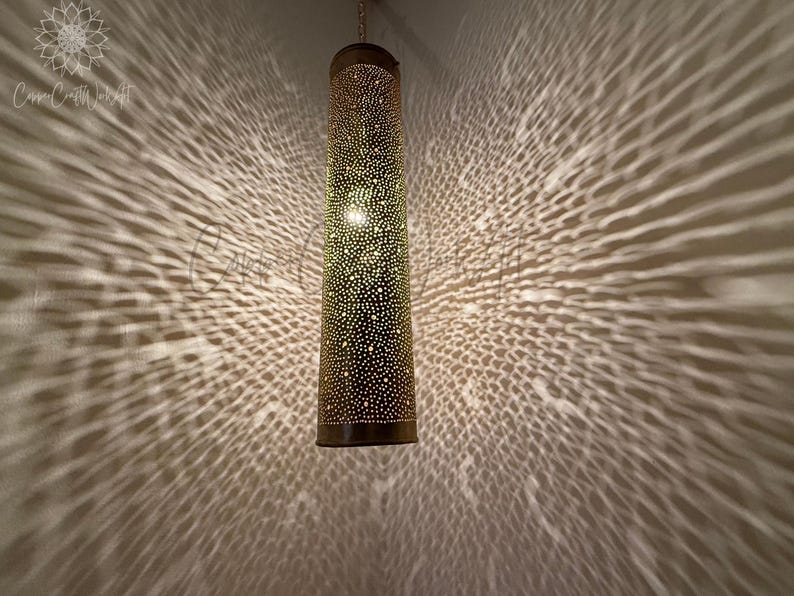 May include: A gold metal hanging lamp with a cylindrical shade featuring a decorative pattern of small holes. The lamp casts a patterned shadow on the wall.