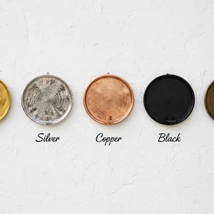 May include: Five circular wall-mounted candle holders arranged in a horizontal row. The finishes are gold, silver, copper, black, and bronze. Each holder features a textured, circular backplate with a small cylindrical candle cup at the base.