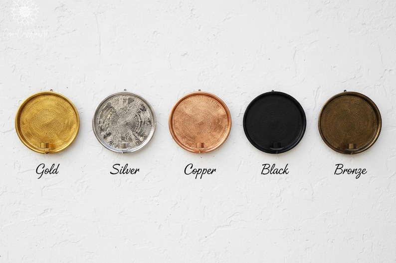 May include: Five circular wall-mounted candle holders arranged in a horizontal row. The finishes are gold, silver, copper, black, and bronze. Each holder features a textured, circular backplate with a small cylindrical candle cup at the base.