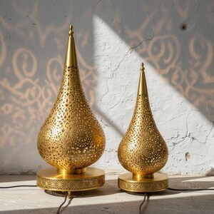 May include: Two gold-coloured metal lamps with a teardrop shape and intricate cut-out patterns. Each lamp features a pointed top and sits on a circular base with matching decorative perforations. The lamps are connected by black electrical cords.