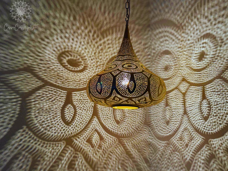 May include: A gold pendant light fixture with a geometric pattern and intricate details. The light casts a decorative shadow on the wall.