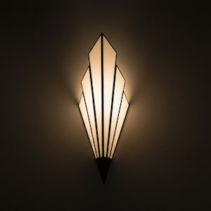 May include: A wall-mounted light fixture featuring a fan-shaped design with multiple vertical panels of frosted white glass. The panels are framed by dark, thin lines that converge at a pointed base, emitting a warm, soft glow from within.