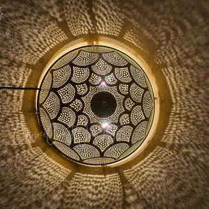 May include: A decorative, circular pendant light with a detailed, perforated metal shade. The shade features a symmetrical pattern of dots and curved shapes, casting intricate shadows on the surrounding wall. The light fixture is suspended from the ceiling.