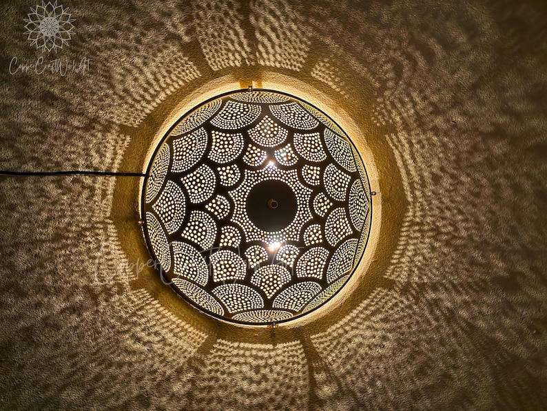 May include: A decorative, circular pendant light with a detailed, perforated metal shade. The shade features a symmetrical pattern of dots and curved shapes, casting intricate shadows on the surrounding wall. The light fixture is suspended from the ceiling.