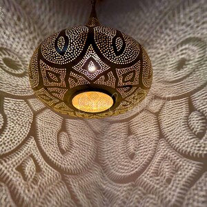 May include: A gold metal Moroccan pendant light with intricate geometric patterns. The light casts a beautiful pattern of light and shadow on the ceiling.