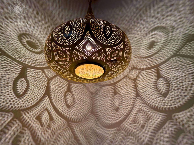 May include: A gold metal Moroccan pendant light with intricate geometric patterns. The light casts a beautiful pattern of light and shadow on the ceiling.