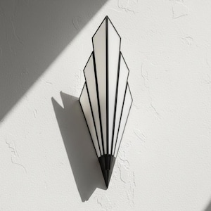 May include: A wall-mounted light fixture featuring a fan-shaped design with five vertical white glass panels framed in black metal. The fixture tapers to a pointed black metal base at the bottom, creating a symmetrical, elongated geometric silhouette.