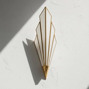 May include: A wall sconce featuring a vertical, fan-shaped design with white glass panels framed in polished gold metal. The fixture tapers to a pointed gold base at the bottom, creating a symmetrical, multi-tiered silhouette that extends outward.