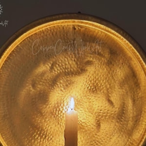 May include: A circular, hammered gold-toned metal wall plaque with a reflective surface. A single white pillar candle stands in the foreground, casting a warm glow onto the textured metal. The text CopperCraftWorkArt is visible on the surface.