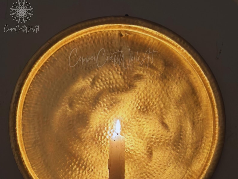 May include: A circular, hammered gold-toned metal wall plaque with a reflective surface. A single white pillar candle stands in the foreground, casting a warm glow onto the textured metal. The text CopperCraftWorkArt is visible on the surface.