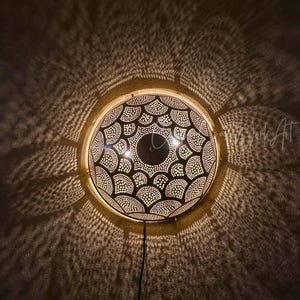 May include: A round, decorative wall lamp with a patterned design. The lamp is made of metal and has a series of small holes that allow light to shine through, creating a pattern on the wall. The lamp is lit from within, casting intricate shadows.