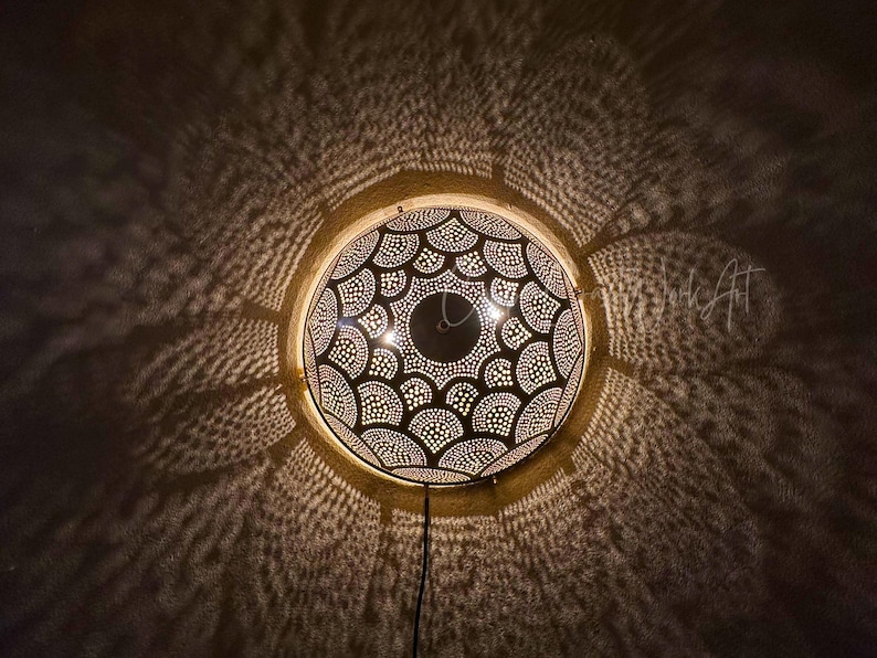 May include: A round, decorative wall lamp with a patterned design. The lamp is made of metal and has a series of small holes that allow light to shine through, creating a pattern on the wall. The lamp is lit from within, casting intricate shadows.