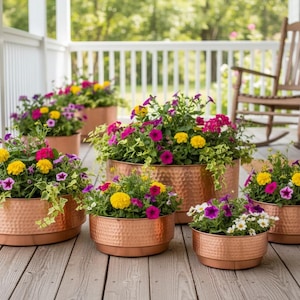 Peut inclure: Un ensemble de cinq jardinières rondes en cuivre martelé de tailles variées, chacune remplie d'un arrangement vibrant de fleurs jaunes, roses et violettes avec un feuillage vert, disposées sur un plancher de porche en bois.