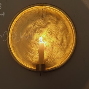May include: A circular gold-toned metal wall sconce featuring a hammered texture and a central candle holder. A single white taper candle is lit, casting a warm glow against the reflective, textured surface of the round metal backplate.