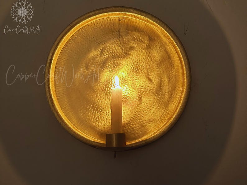 May include: A circular gold-toned metal wall sconce featuring a hammered texture and a central candle holder. A single white taper candle is lit, casting a warm glow against the reflective, textured surface of the round metal backplate.