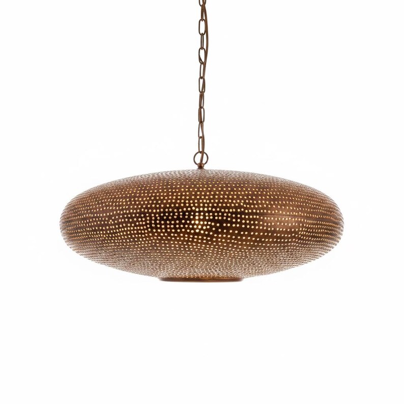 May include: A round, flattened pendant light fixture with a metallic bronze finish. The surface is covered in a dense pattern of small, circular perforations that allow light to pass through, suspended from the ceiling by a matching metal chain.