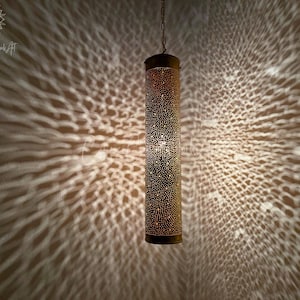 May include: A gold metal pendant light fixture with a cylindrical shade featuring a decorative pattern of small holes. The light casts a geometric pattern on the wall behind it.