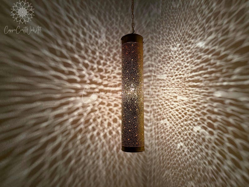May include: A gold metal pendant light fixture with a cylindrical shade featuring a decorative pattern of small holes. The light casts a geometric pattern on the wall behind it.