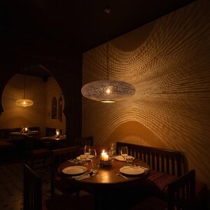 May include: A metallic, oval-shaped pendant light fixture with a perforated surface that casts intricate, radiating light patterns onto the surrounding walls. The lamp hangs from a thin chain above a circular wooden dining table set with plates.