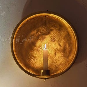 May include: A circular, hammered gold-toned metal wall sconce featuring a central candle holder. The reflective, textured surface of the round backplate catches the light from a single white pillar candle positioned in the center of the fixture.