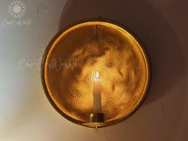 May include: A circular, hammered gold-toned metal wall sconce featuring a central candle holder. The reflective, textured surface of the round backplate catches the light from a single white pillar candle positioned in the center of the fixture.