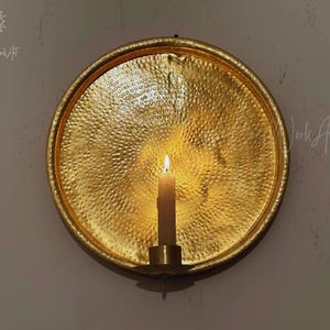 May include: A circular gold-toned metal wall sconce featuring a hammered, textured surface. A single cylindrical candle holder is attached to the center, holding a lit off-white taper candle that casts a warm glow across the reflective metal plate.