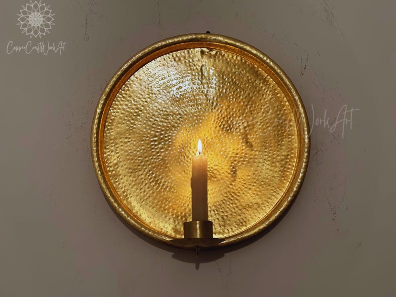 May include: A circular gold-toned metal wall sconce featuring a hammered, textured surface. A single cylindrical candle holder is attached to the center, holding a lit off-white taper candle that casts a warm glow across the reflective metal plate.