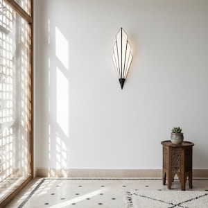 May include: A wall-mounted sconce featuring a geometric, fan-like shape with black metal framing and white panels. The light fixture emits a soft glow, mounted on a plain white wall above a small, carved wooden side table holding a potted succulent.