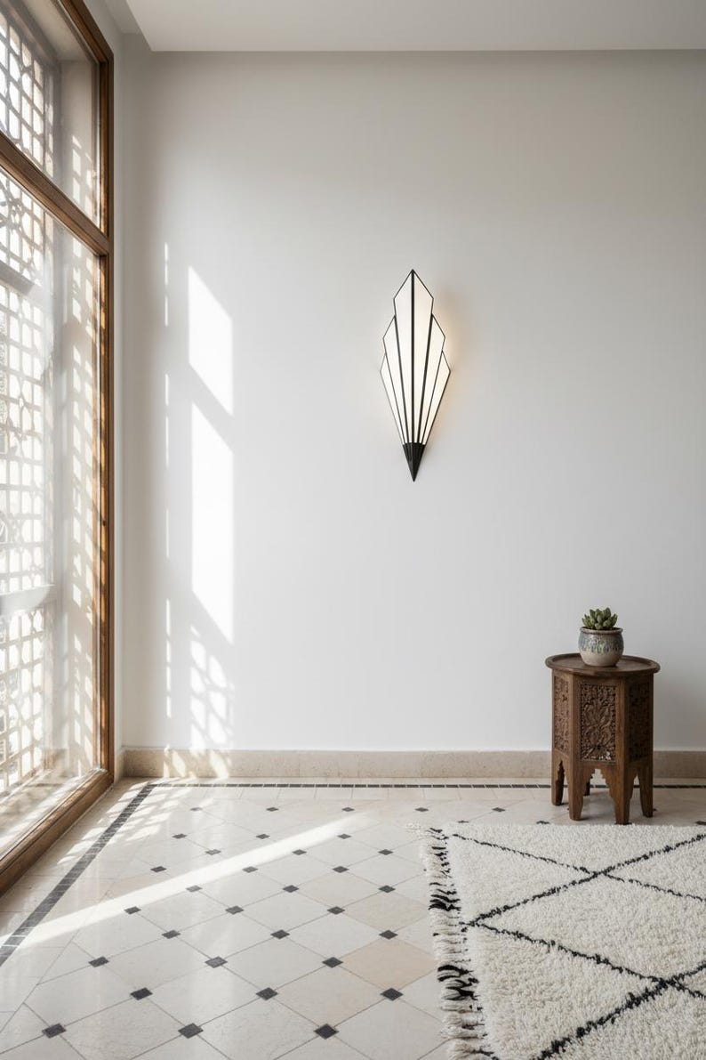 May include: A wall-mounted sconce featuring a geometric, fan-like shape with black metal framing and white panels. The light fixture emits a soft glow, mounted on a plain white wall above a small, carved wooden side table holding a potted succulent.